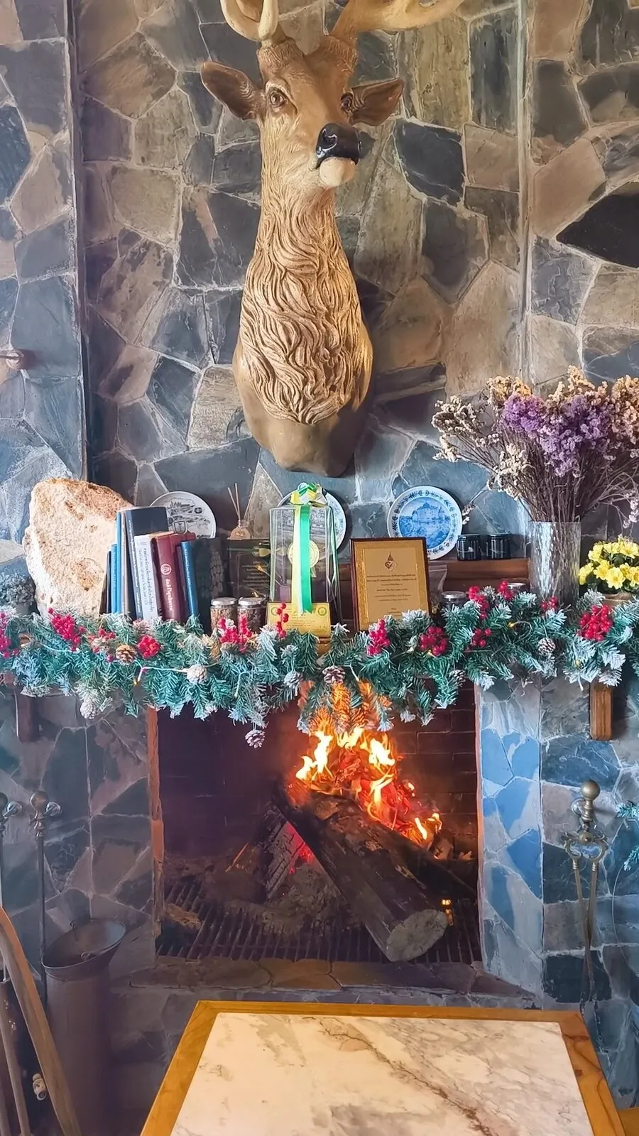A fire burning in a stone fireplace beneath a mounted deer head and Christmas garland