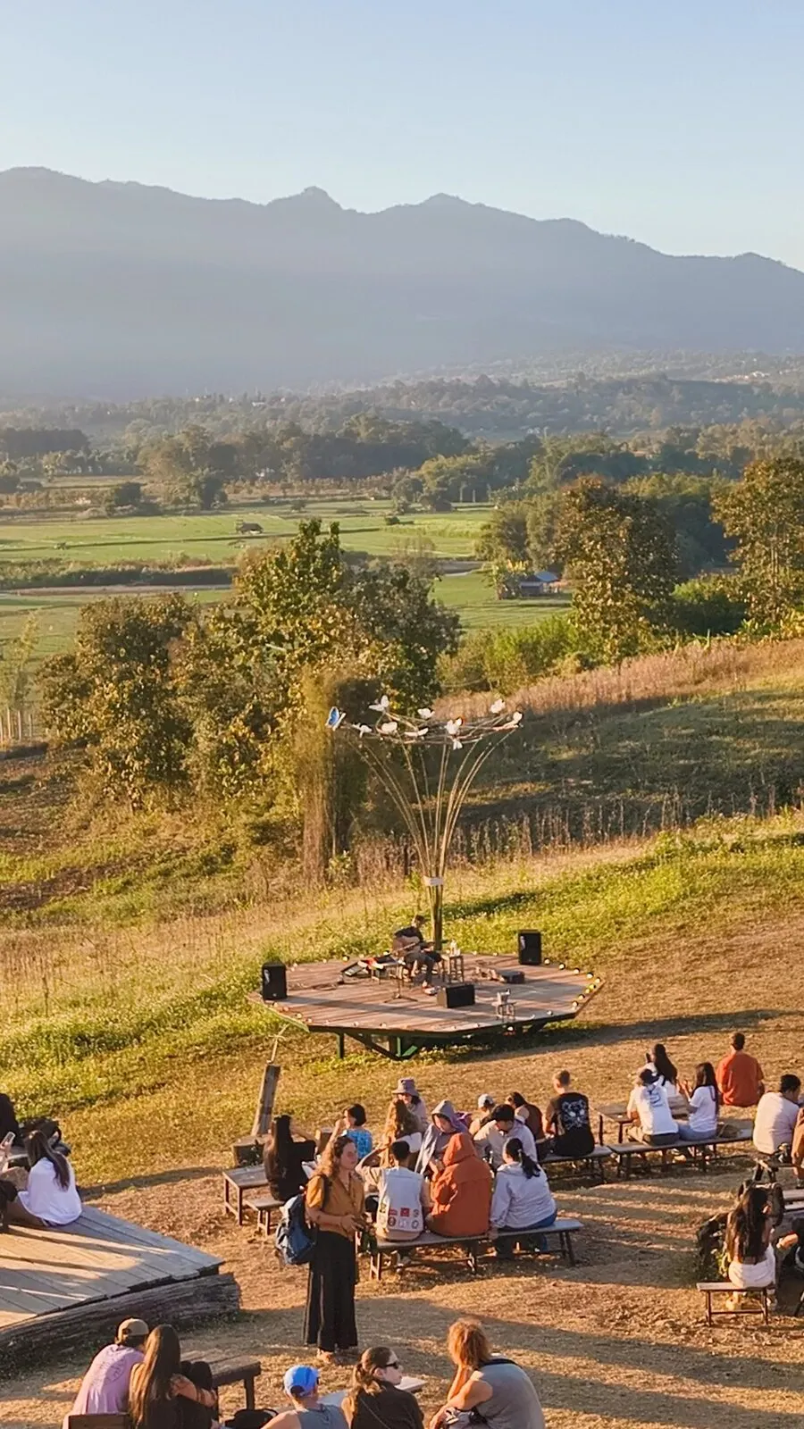 A crowd gathered on a grassy field in front of a small stage at golden hour