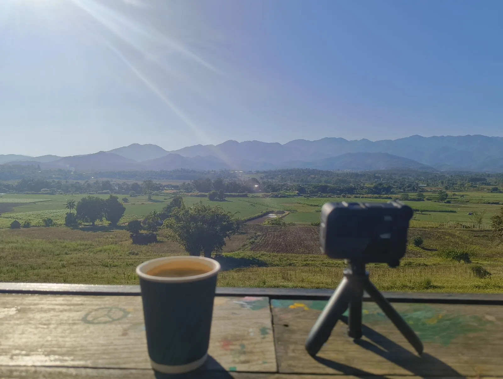A coffee cup and a small tripod on a wooden terrace overlooking green fields