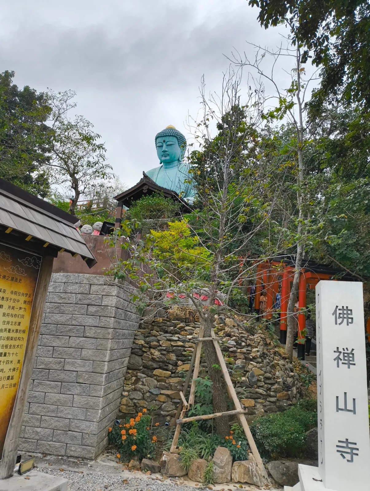 The teal Buddha rising above trees beside a white stone pillar with Chinese characters