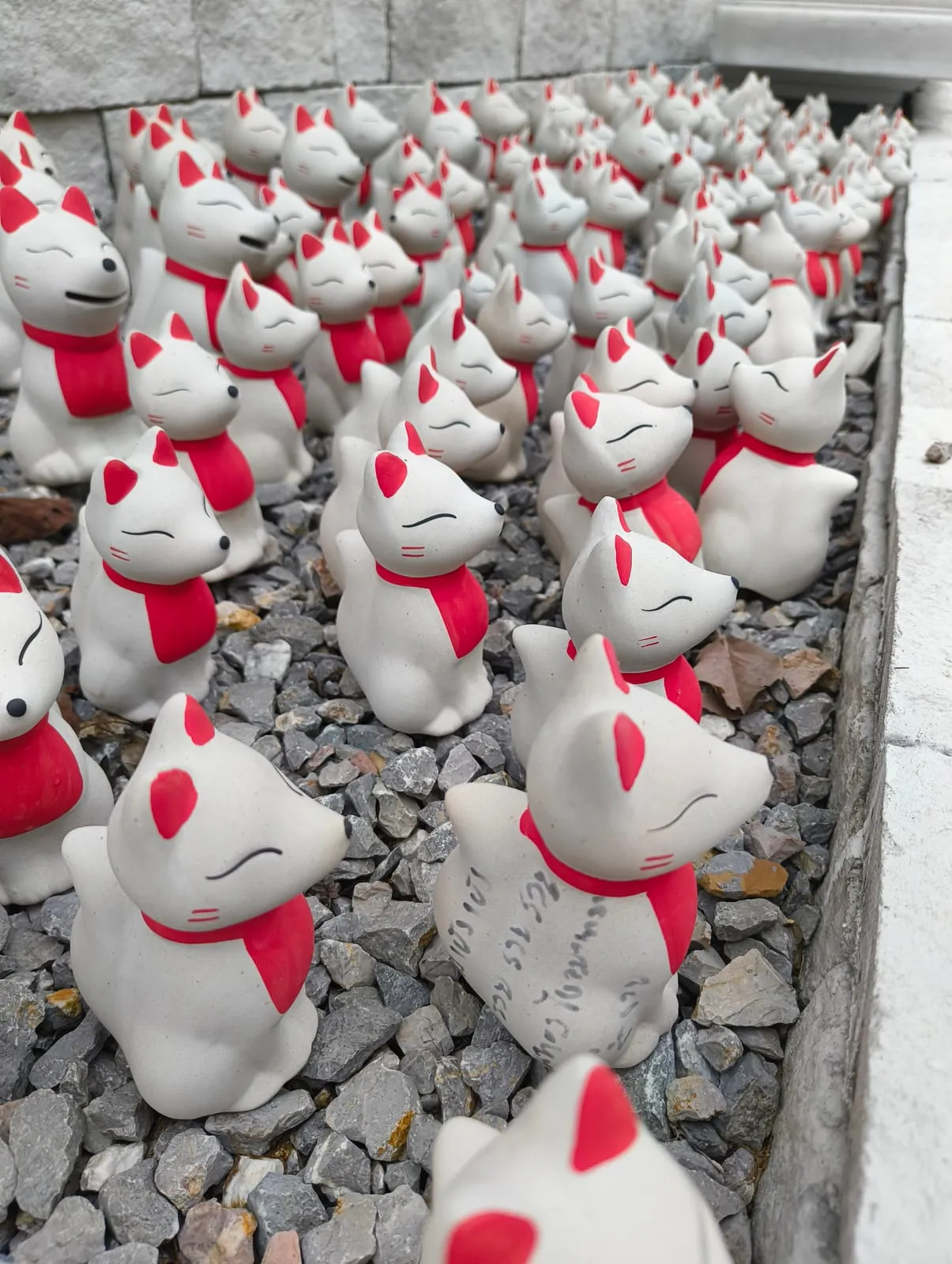 A crowd of small white kitsune statues with red bibs on grey gravel