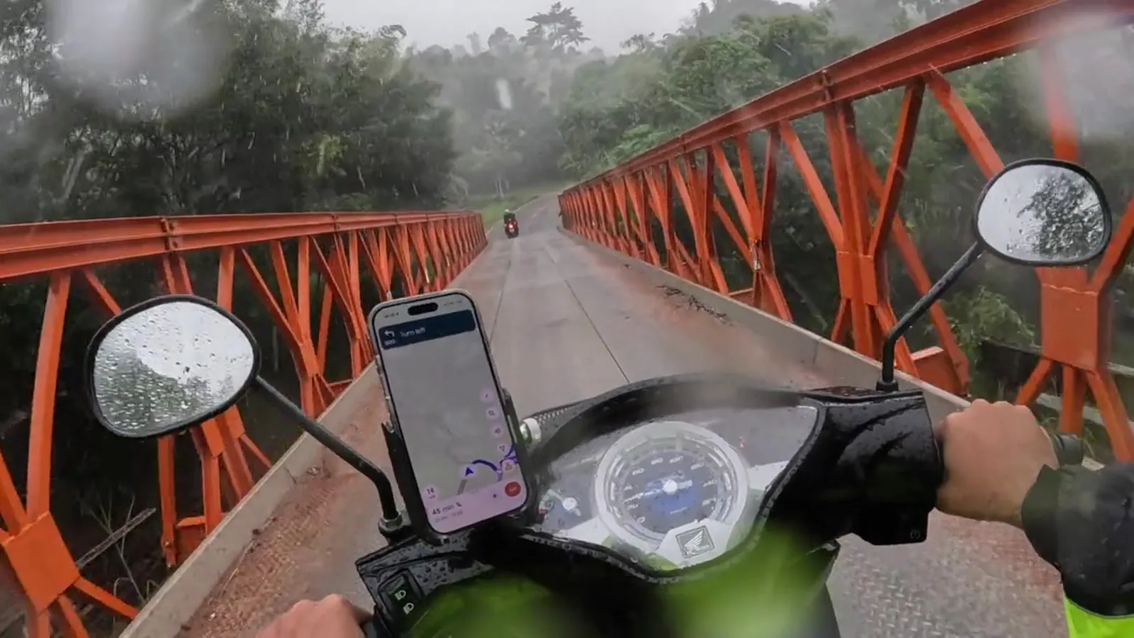 A rider crossing a long orange truss bridge in the rain, following another rider