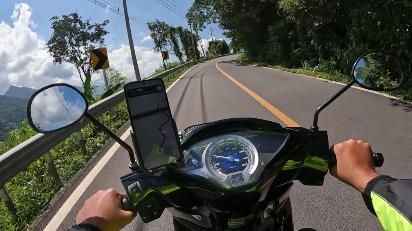 A rider looking over the handlebars at a sunlit road curving between forested ridges, a yellow chevron sign ahead