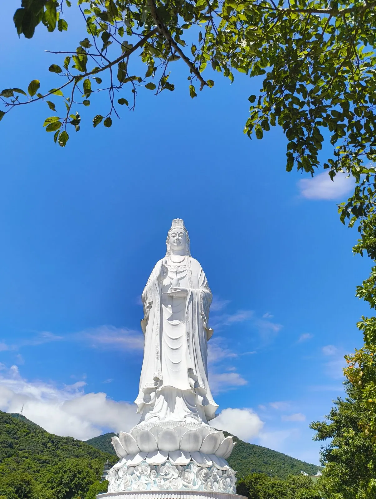 A tall white Lady Buddha statue standing above the trees, with mountains behind